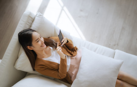 High angle view of happy cheerful positive young Asian woman on the phone at home, smiling girl using app and browsing on the internet on smartphone lying on sofaの写真素材