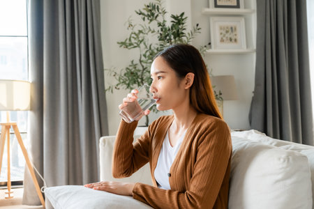 Happy young Asian woman drinking water while sitting on the sofa in the living roomの写真素材