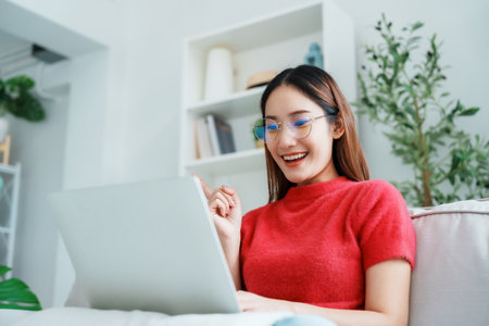 Happy young Asian woman using laptop while seated on couch at homeの写真素材