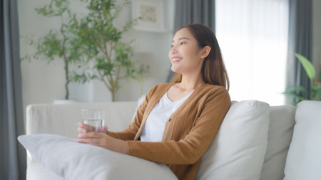 Happy young Asian woman drinking water while sitting on the sofa in the living roomの写真素材