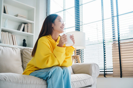 Young asian woman wearing eyeglass sitting on sofa and holding mug in living room at home, drinking milk or tea in the morning. Healthy and refresh moodの写真素材