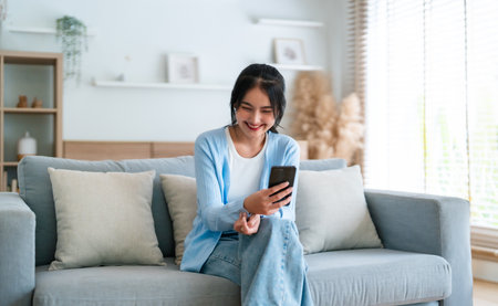 Smiling pretty young Asian woman sitting on cozy couch, using funny mobile apps in living room. Woman at Home, Doing Online Shopping, Messaging Friends, Posting on Social Mediaの写真素材