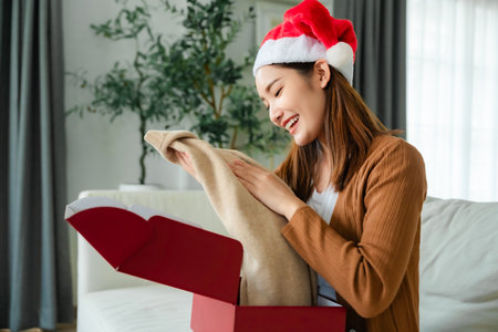 Young Asian woman wearing Christmas hat and opening a gift box, sitting on sofa in living room. Happy new year, Thanksgiving day, Merry Christmasの写真素材