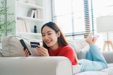 Young asian woman lying on sofa in living room, makes online banking payments through the internet from bank card on cell phone. Shopping online on mobile with credit cardの写真素材