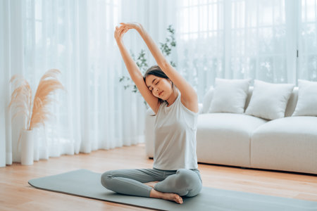Happy young Asian woman practicing yoga and shoulder stretching at home sitting on floor in living roomの写真素材