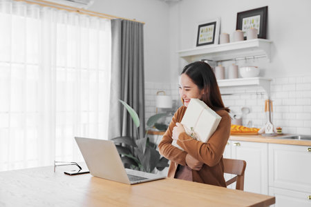Young asian woman using computer laptop at home. Female showing gift box while on video conversation with friend. Happy birthday, Happy new year, Thanksgivingの写真素材