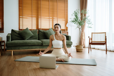 Young Asian woman meditating in lotus position at home, sitting on the floor in fitness clothing. Breathing workout to achieve relaxation, peace and mindfulnessの写真素材