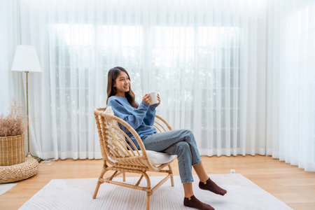 Young Asian woman happy and smiling, sitting on seat and holding mug in living room at homeの写真素材