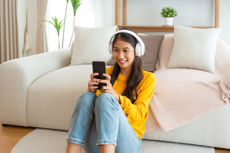 Happy young asian woman relaxing at home. Female smiling and wearing headset sitting on the floor beside sofa and holding mobile smartphone. Girl using video call to close friendの写真素材