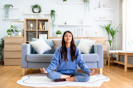 Happy young Asian woman practicing yoga and meditation at home sitting on floor in living room in lotus position and relaxing with closed eyes. Mindful meditation and wellbeing conceptの写真素材