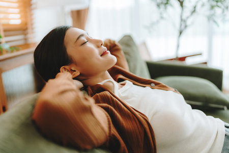 Relaxed young Asian woman enjoying rest on comfortable sofa, calm attractive girl relaxing on couch, breathing fresh air with eyes closed, meditating at homeの写真素材