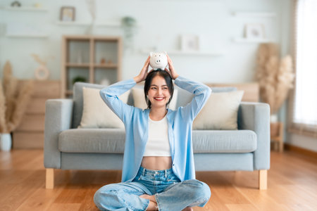 Young Asian woman holding piggy bank and sitting on the floor at home. Save money and financial investmentの写真素材