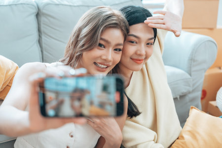 Two young Asian women smiling and happy while taking a selfie in their living room at home, looking at the cameraの写真素材