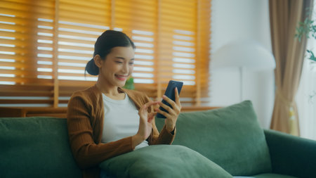 Smiling pretty young Asian woman sitting on cozy couch, using funny mobile apps in living room. Woman at home, Doing online shopping, Messaging friends, Posting on social Mediaの写真素材