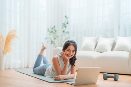 Young Asian woman lying on the floor on yoga mat in front of laptop and training at home. Beautiful smiling woman doing stretching exercise while communicates with a trainer online before workoutの写真素材
