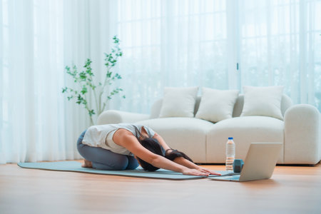 Young Asian woman stretching yoga workout on exercise mat while online training class with computer laptop in living roomの写真素材