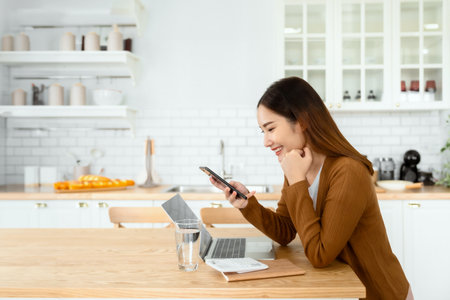 Happy young asian woman relax in domestic kitchen at home, using smartphone, smiling girl use cell phone chatting, browse wireless internet on gadget, shopping online from homeの写真素材