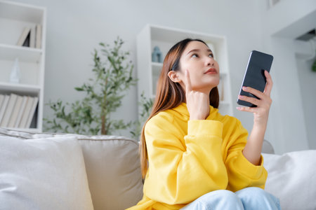 Happy young asian woman relax on comfortable couch at home texting messaging on smartphone, smiling girl use cellphone chatting, browse wireless internet on gadget, shopping online from homeの写真素材