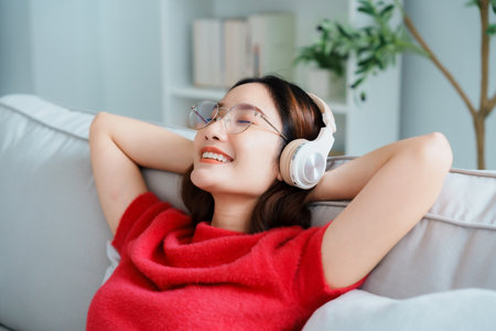 Relaxed young Asian woman enjoying rest on comfortable sofa, calm attractive girl relaxing on couch, breathing fresh air with eyes closed, wearing eyeglasses and headset, meditating at homeの写真素材