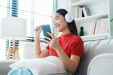 Young asian woman listening to music on couch in living room at home. Happy asia female using mobile smartphone, wearing headset and sitting on sofaの写真素材