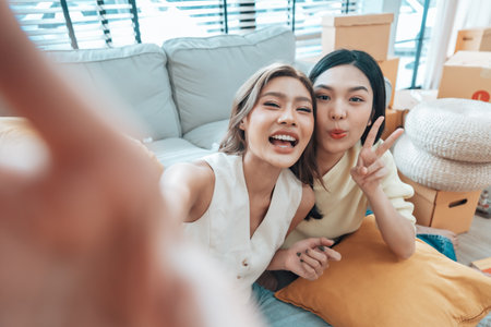Two young Asian women smiling and happy while taking a selfie in their living room at home, looking at the cameraの写真素材
