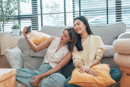 Two young Asian women smiling and happy while taking a selfie in their living room at home, looking at the cameraの写真素材
