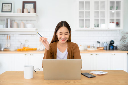 Beautiful young Asian woman working with laptop computer in home kitchen. Work at home. Remote studying, Virtual training, E-learning, Watching online education, Webinar at houseの写真素材