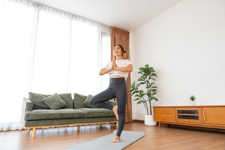 Young beautiful Asian woman practices tree pose during a morning yoga session in living room at homeの写真素材