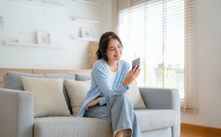 Smiling pretty young Asian woman sitting on cozy couch, using funny mobile apps in living room. Woman at Home, Doing Online Shopping, Messaging Friends, Posting on Social Mediaの写真素材