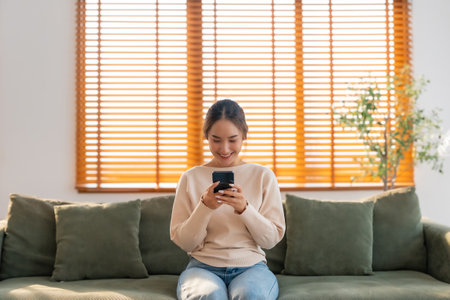 Happy young asian woman relax on comfortable couch at home texting messaging on smartphone, smiling girl use cellphone, chatting online message, shopping online from homeの写真素材