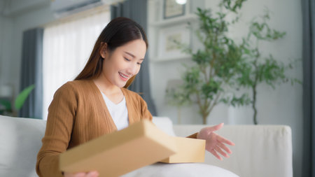 Happy young Asian woman opening delivery package at home. Beautiful female customer open box and sitting on couch in living roomの写真素材