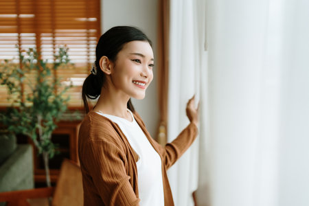 Happy Asian woman standing by the window and looking outside the living room enjoying the bright sunshineの写真素材
