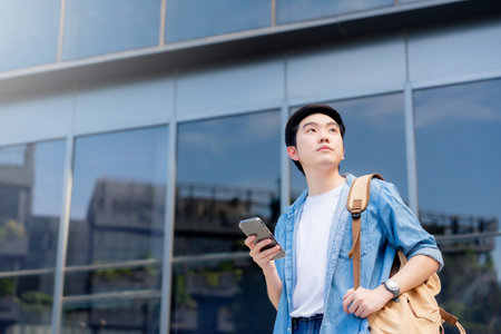 Portrait of handsome Asian student using smartphone. A young man standing outdoor happy smiling with holding mobile phoneの写真素材
