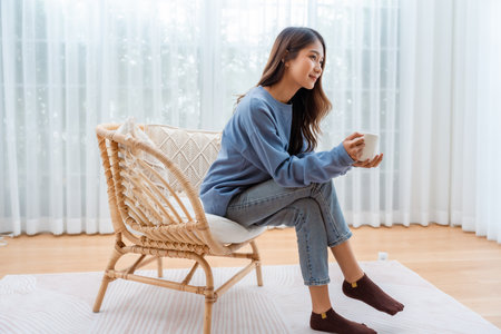 Young Asian woman happy and smiling, sitting on seat and holding mug in living room at homeの写真素材