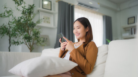 Happy young asian woman relax on comfortable couch at home texting messaging on smartphone, smiling girl use cellphone, chatting online message, shopping online from homeの写真素材