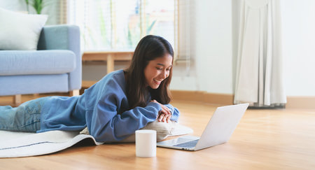 Young asian woman smile working in living room at home. Happy female using computer laptop and lying on the floor at house, Video call with friendの写真素材