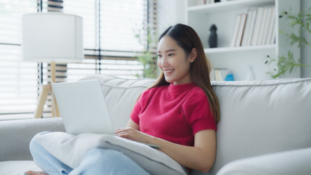Happy young Asian woman using laptop while seated on couch at homeの写真素材
