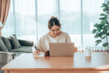 Happy young Asian woman working on laptop in living room. Freelance worker female sitting at desk writing notes while watching webinar, studying onlineの写真素材