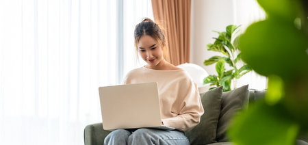 Happy young Asian woman working on laptop in living room. Freelance worker female sitting couch while watching webinar, studying onlineの写真素材