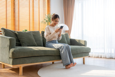 Happy young asian woman relax on comfortable couch at home texting messaging on smartphone, smiling girl use cellphone, chatting online message, shopping online from homeの写真素材