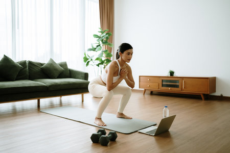 Young Asian beautiful woman doing aerobic exercise at home. Attractive girl doing  workout by squatting, follow instructions video from online trainer for health in houseの写真素材