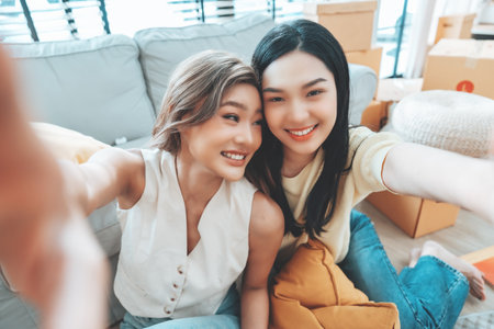 Two young Asian women smiling and happy while taking a selfie in their living room at home, looking at the cameraの写真素材