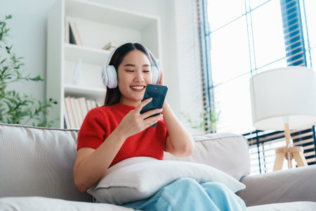 Young Asian woman relaxing on sofa at home, listening music with headset on cell phone. People lifestyle conceptの写真素材