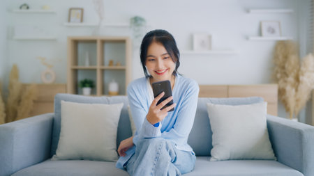 Smiling pretty young Asian woman sitting on cozy couch, using funny mobile apps in living room. Woman at Home, Doing Online Shopping, Messaging Friends, Posting on Social Mediaの写真素材