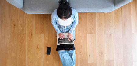 Top view of young Asian woman sitting on the floor and working on computer laptop with mobile smart phone in living room at homeの写真素材