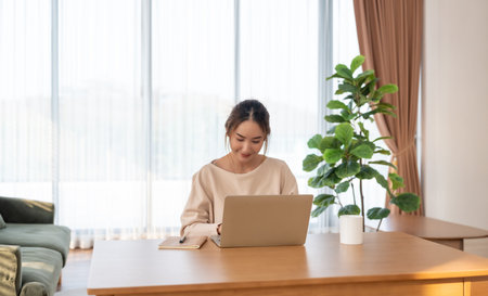 Happy young Asian woman working on laptop in living room. Freelance worker female sitting at desk writing notes while watching webinar, studying onlineの写真素材