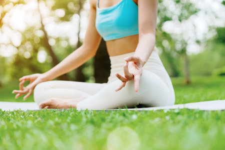 Asian woman meditating in a peaceful park setting, sitting cross-legged on green grass with trees in the background on a sunny day. Concept of mindfulness, self-care, and outdoor relaxationの写真素材