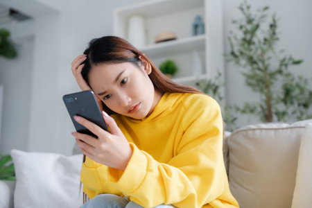 News on device screen. Upset asian woman frustrated by problem with work or relationships, sitting on couch, feeling despair and anxiety, loneliness, having psychological troubleの写真素材