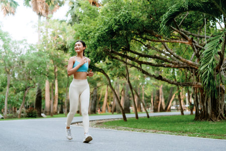 Asian woman jogging on a pathway in a public park, wearing athletic clothing and enjoying a cardio workout in a green, natural environmentの写真素材
