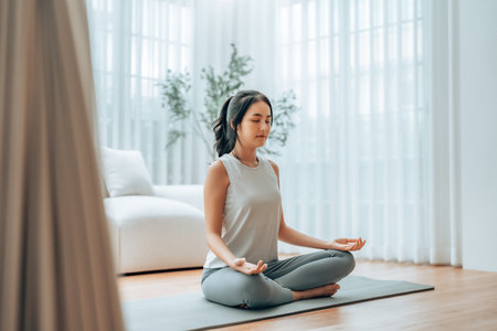 Young Asian woman meditating in lotus position at home, sitting on the floor in fitness clothing. Breathing workout to achieve relaxation, peace and mindfulnessの写真素材